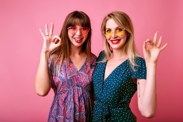 Two happy best friends sister women girls wearing trendy spring summer printed bright vintage dresses and sunglasses , hugs and making ok science gesture by their hands.