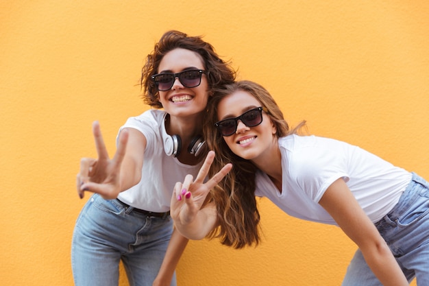 Two happy cheerful teenage girls in sunglasses showing peace gesture