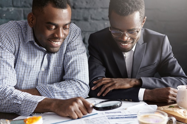 Free photo two happy dark-skinned businessmen reading documents with magnifying glass