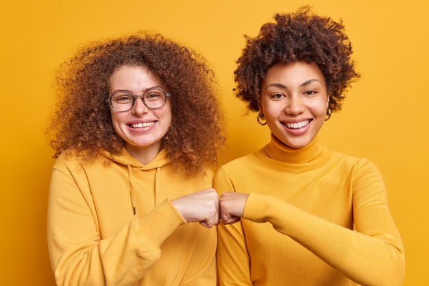 Two happy diverse women make fist bumps demonstrate agreement have friendly relationship smile gladfully stand next to each other isolated over yellow wall. Teamwork body language concept