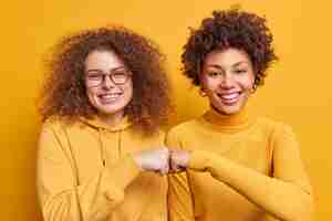 Бесплатное фото Две счастливые разноплановые женщины хлопают кулаками, демонстрируя согласие в дружеских отношениях, улыбаются, радостно стоять рядом друг с другом, изолированными над желтой стеной. концепция языка тела совместной работы