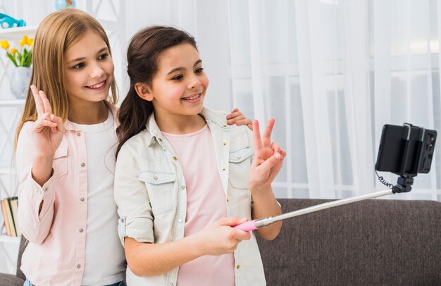 Two happy female friend making peace gesture taking selfie on smartphone at home