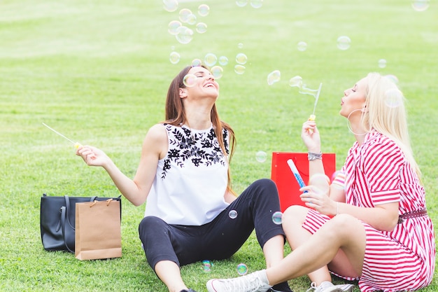 Free photo two happy female friends sitting on green grass blowing bubbles