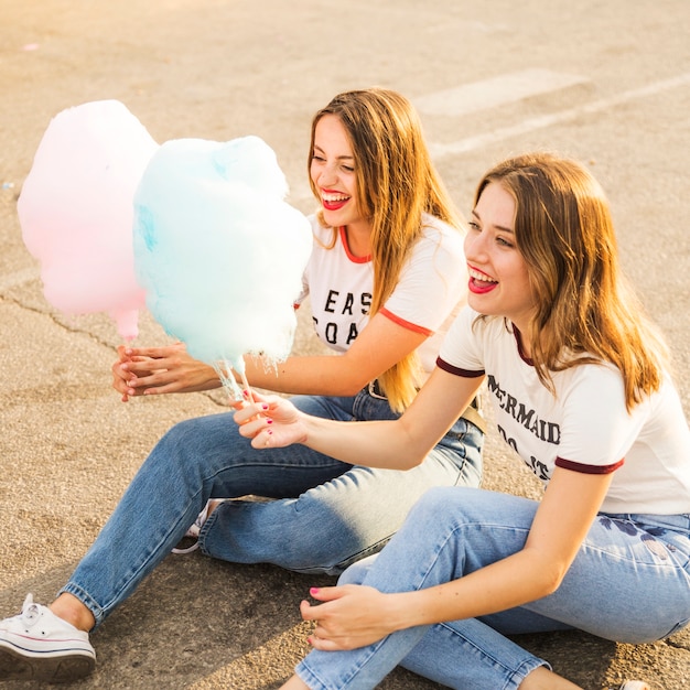 Two happy female friends sitting on street holding candy floss