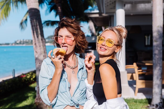 분홍색과 노란색 선글라스에 두 행복 맞는 여자 재미 도넛, 야외와 함께 웃고 웃고