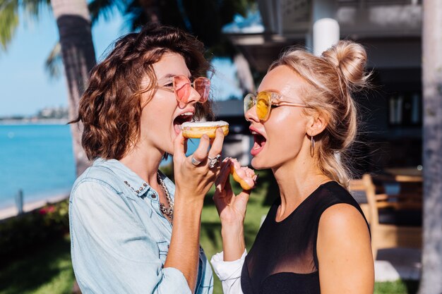 Two happy fit woman in pink and yellow sunglasses smiling having fun laughing with donuts, outdoor