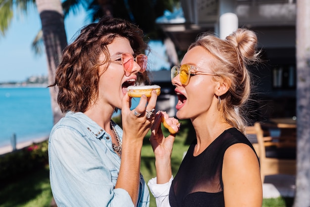 Две счастливые женщины в розовых и желтых солнцезащитных очках улыбаются, весело смеются с пончиками, на открытом воздухе