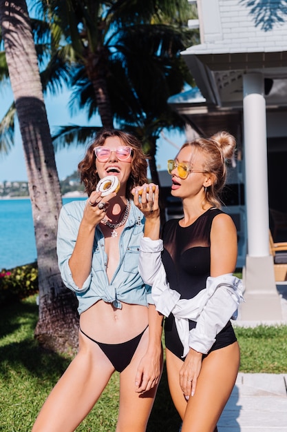 Free photo two happy fit woman in pink and yellow sunglasses smiling having fun laughing with donuts, outdoor