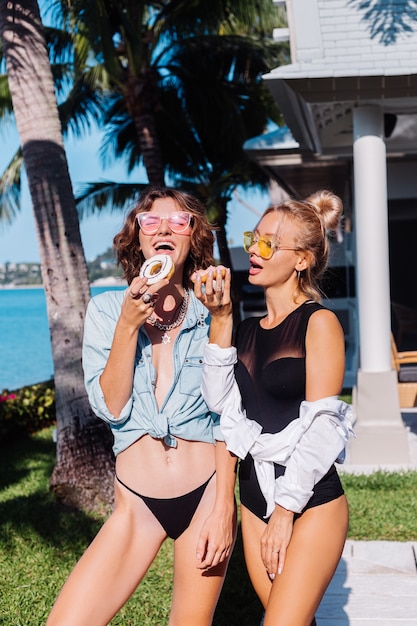 Two happy fit woman in pink and yellow sunglasses smiling having fun laughing with donuts, outdoor