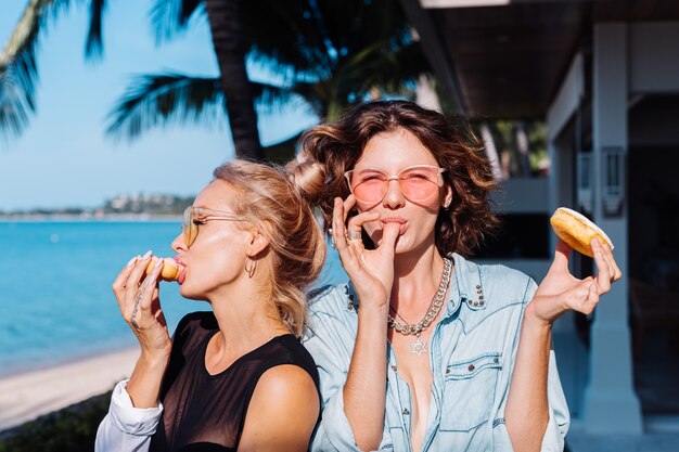 Two happy fit woman in pink and yellow sunglasses smiling having fun laughing with donuts, outdoor