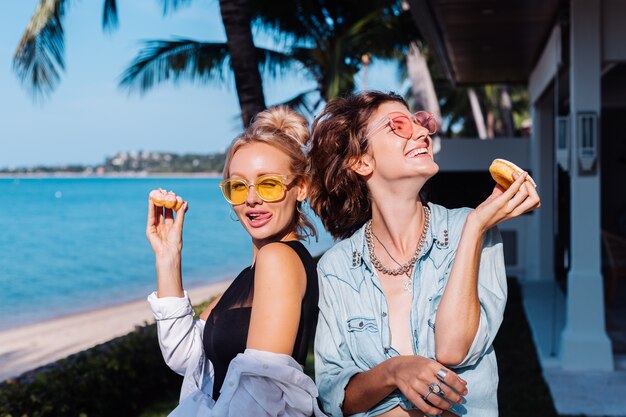 Two happy fit woman in pink and yellow sunglasses smiling having fun laughing with donuts, outdoor