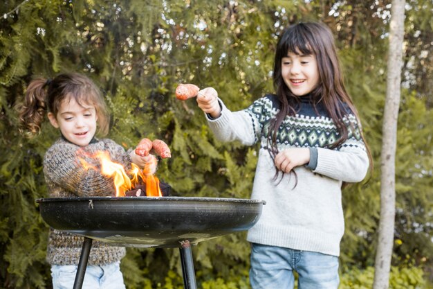 Two happy girls grilling sausages in fire on portable barbecue