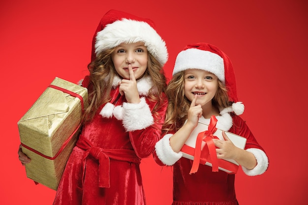 Two happy girls in santa claus hats with gift boxes on red