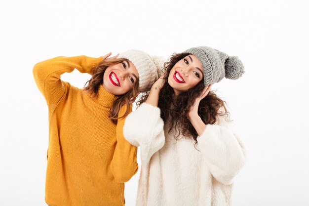 Two happy girls in sweaters and hats having fun together  over white wall