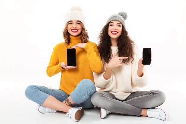 흰 벽 위에 공백 스마트 폰 화면을 표시하면서 함께 바닥에 앉아 스웨터와 모자에 두 행복 소녀