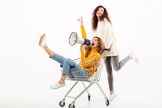 Two happy girls in sweaters having fun with shopping trolley and megaphone over white wall