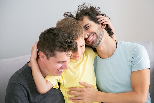 Free photo two happy handsome fathers and son sitting on couch together and hugging each other. happy family and parenthood concept