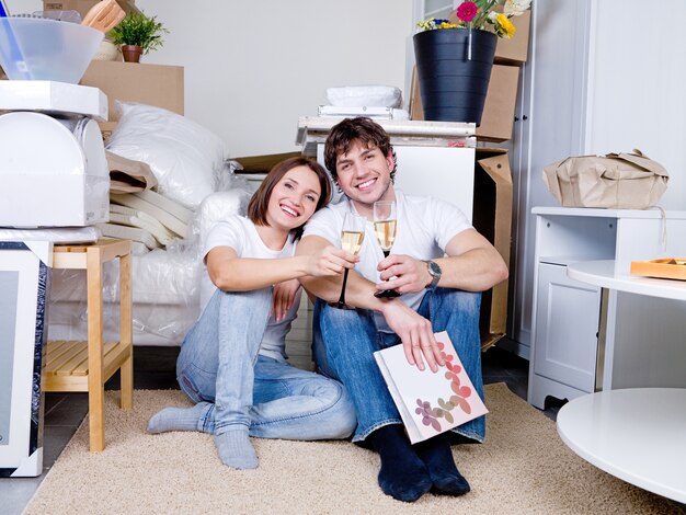 Два счастливых человека сидят на полу в своей новой квартире с бокалом шампанского