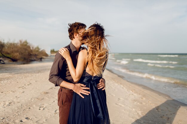 Two happy people in love, elegant woman with her handsome boyfriend hugging and enjoying romantic time together on the beach. Elegant dress.