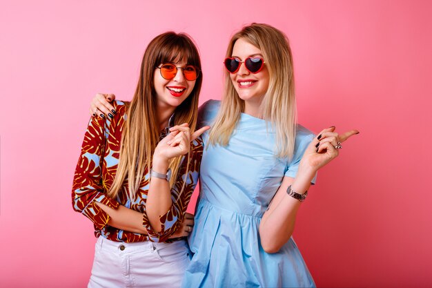 Two happy pretty sisters best friends hipster women having fun together at pink wall, hugs and kisses, happy couple, trendy bright summer clothes and accessories, relationship goals.