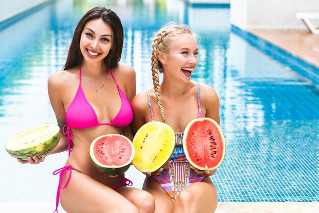 Two happy pretty woman having fun near pool at summer party, holding watermelons and wearing swimsuits