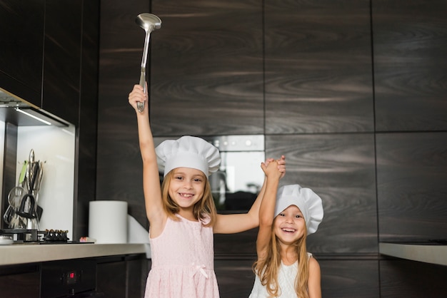 Two happy sisters wearing chef hat in kitchen holding their hands