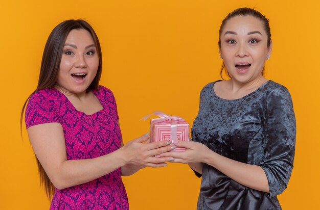 국제 여성의 날을 축하하는 두 명의 행복하고 놀란 아시아 여성이 오렌지 벽 위에 넓게 서있는 선물을 들고
