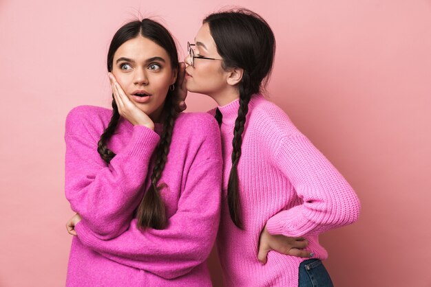 Two happy teenage girls with braids in casual clothes gossiping together isolated over pink wall