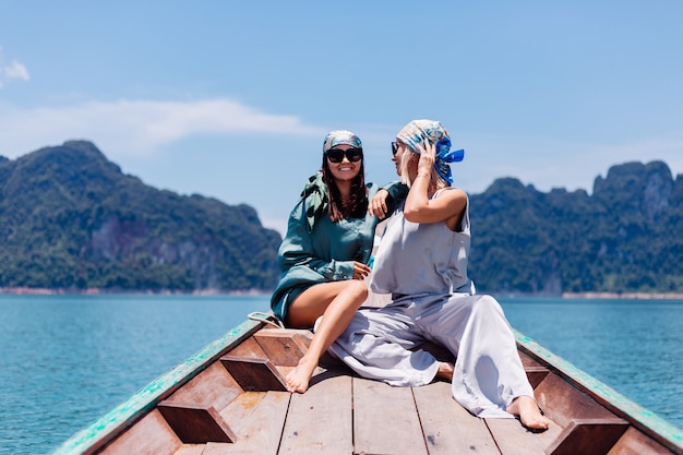 Два счастливых туристических друга-блогера женщины в шелковом костюме, шарфе и солнечных очках во время отпуска путешествуют по Таиланду на азиатской лодке в национальном парке Кхао Сок.