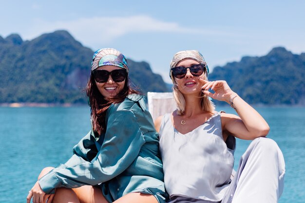 Two happy woman blogger tourist friends in silk suit and scarf and sunglasses on vacation travel around thailand on asian boat, Khao Sok national park.