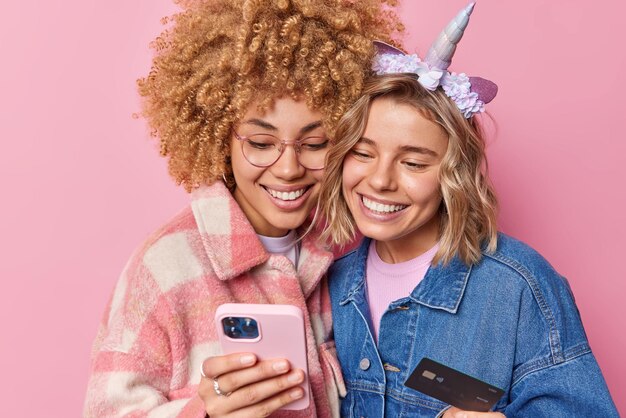 두 명의 행복한 여성이 스마트폰 다운로드 온라인 결제 앱에서 즉시 돈을 이체하여 신용 카드 계정 잔액을 확인하여 인터넷 쇼핑을 분홍색 벽에 격리된 서로 가깝게 만듭니다.