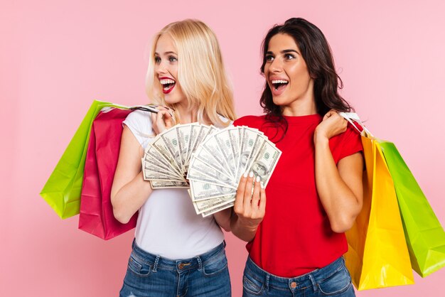 Две счастливые женщины позируют с деньгами и пакетами, глядя на розовый
