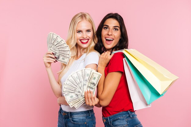 Two happy women posing with money and packages while looking at the camera over pink 