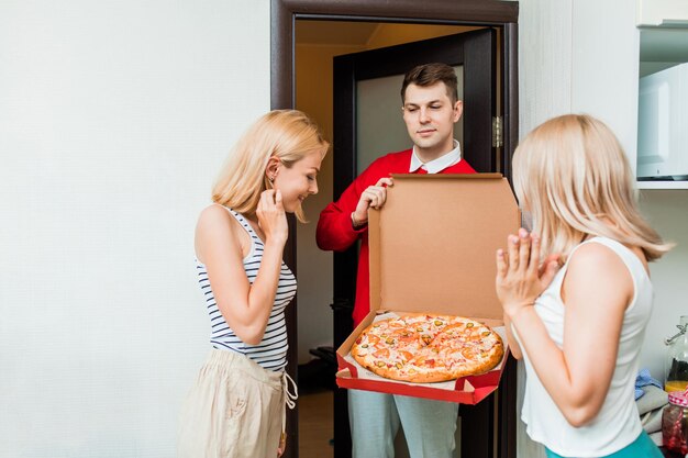 두 명의 행복한 백인 여성이 집에서 배달원에게서 피자를 받고 있습니다. 빨간 제복을 입은 배달원은 문 앞에서 고객에게 피자를 보여줍니다. 음식 배달.