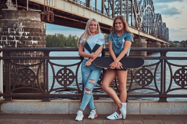 두 명의 행복한 젊은 힙스터 소녀가 스케이트보드를 들고 오래된 다리 배경의 난간에 기대어 있습니다.