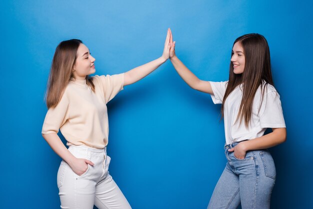 Two happy young women with different hair giving high five to each other isolated over blue wall