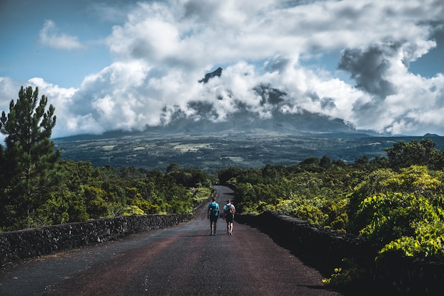 Two hikers walking on a narrow road surrounded with greenery with cloudy mountain