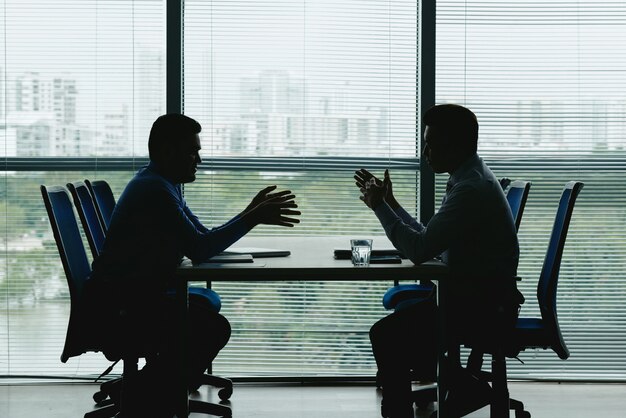 向かい合って座って交渉するシャッター付きのオフィスの窓に対する2つの人間の輪郭