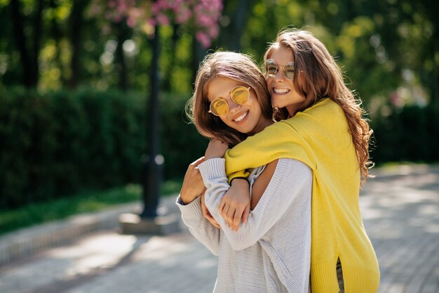 거리에서 포옹하는 밝은 선글라스와 스웨터, 긍정적 인 분위기, 진정한 감정을 가진 두 명의 놀라운 소녀. 거리에서 두 젊은 아가씨의 야외 초상화