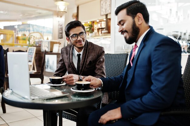 Two indian business man in suits sitting at office on cafe looking at laptop and drinking coffee