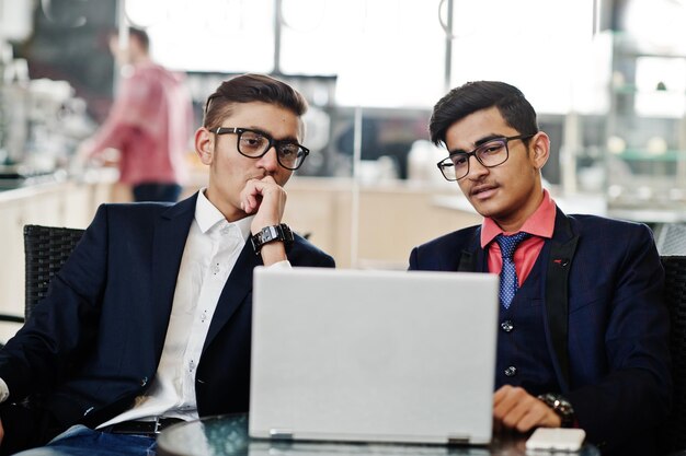 Two indian business man in suits sitting at office on cafe and looking at laptop