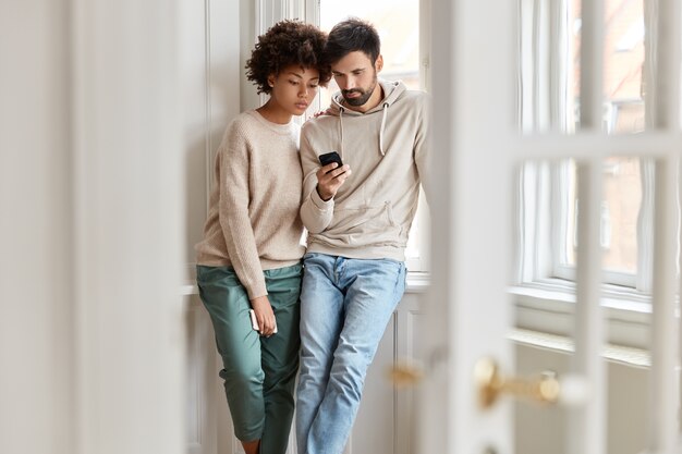 Two interracial students watch attentively tutorial video browsed on modern cell phone, learn course online, pose against domestic view near window, connected to 4G internet, read information