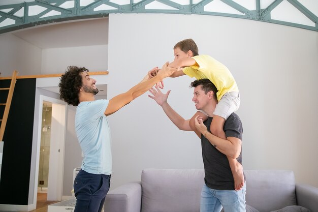 Two joyful dads and son having fun at home, boy riding on mans neck. Side view. Family and parenthood concept