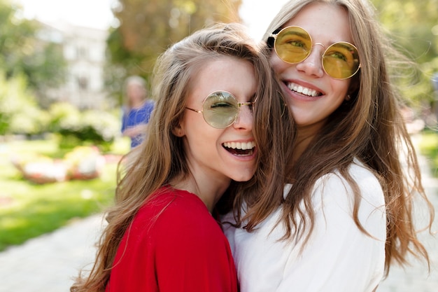 Two joyful pretty girls having fun outdoor in sunny morning on city-view. Stylish fashionable girls in bright sunglasses hugging. Happy time, fashion, relaxation