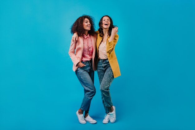 Two joyful women posing on blue studio wall