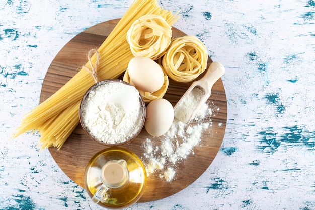 Two kinds raw pasta with oil, egg and bowl of flour on wooden board.