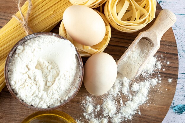 Two kinds raw pasta with oil, egg and bowl of flour on wooden board.