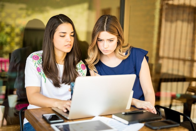 Two latin women sitting at a cafe table with laptop computer and discussing work