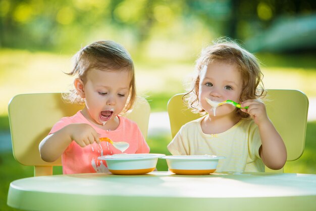 Two little 2 years old girls sitting at a table and eating together against a green lawn