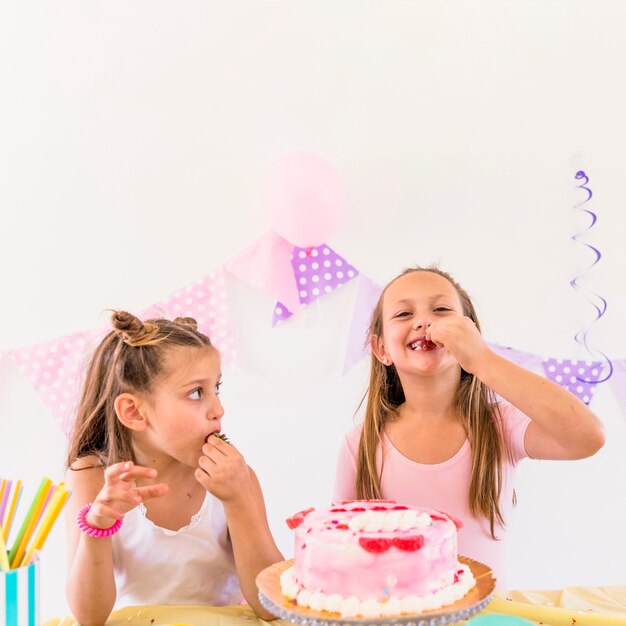 Two little girls eating strawberries in front of delicious cake on table